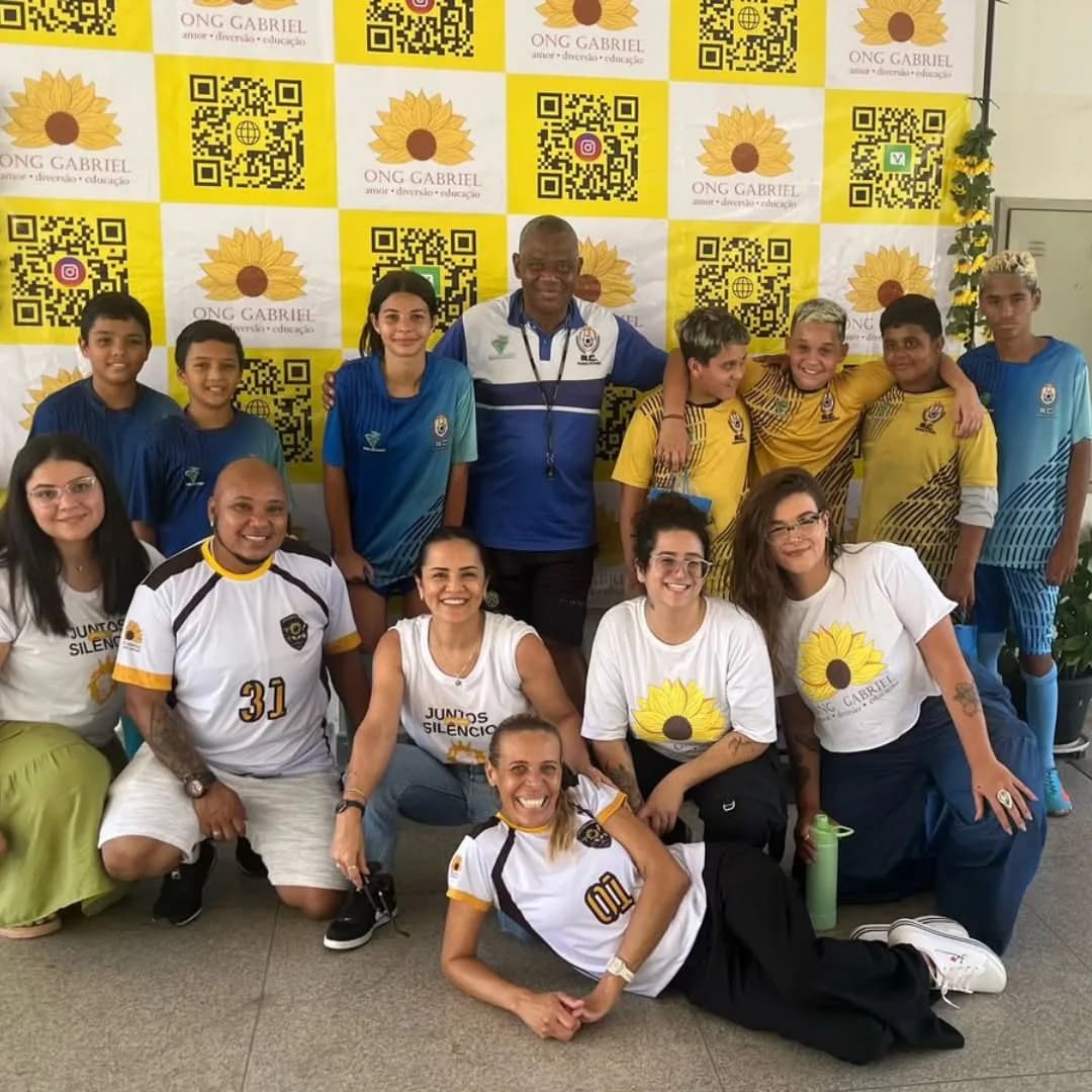 Grupo de crianças, jovens e adultos da ONG Gabriel posando diante de um painel com a marca e códigos QR.