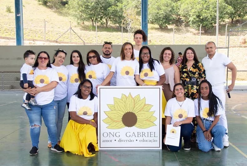 Pessoas da ONG Gabriel reunidas em uma quadra. No centro, duas pessoas seguram um cartaz com o girassol da organização e as palavras amor, diversão e educação.